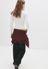 Bílé tričko s dlouhým rukávem v kombinaci s vínovou zavinovací sukní, která má elastický pas, splývavý design a volné černé kalhoty.
