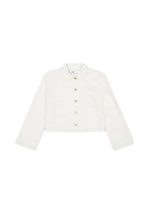 Frau mit langen braunen Haaren trägt eine weiße bauchfreie Jacke und hellblaue Jeans, seitlich vor einem schlichten weißen Hintergrund.