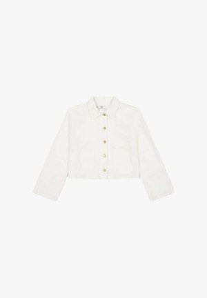 Vrouw met lang bruin haar draagt een witte cropped jas en lichtblauwe spijkerbroek, zijwaarts gezien tegen een effen witte achtergrond.