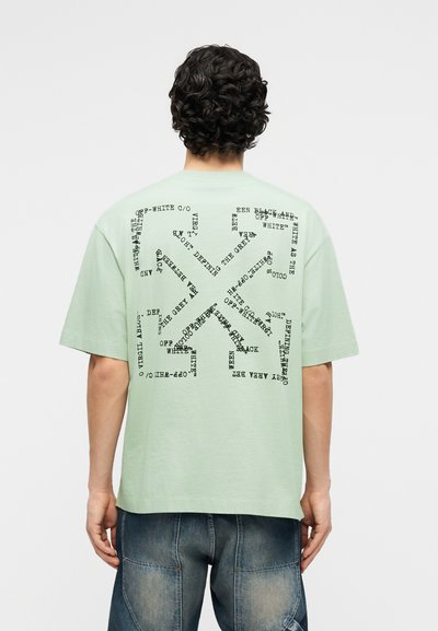 Personne aux cheveux bouclés porte un T-shirt vert clair avec un texte noir formant un motif abstrait dans le dos, associé à un short en jean bleu.