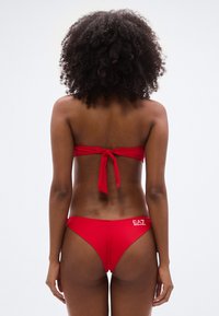 Κόκκινο μπικίνι με  τοπ μπαντό που έχει λεπτομέρεια δέσιμου στο πίσω μέρος. Το κάτω μέρος είναι σε χαμηλή μέση με λογότυπο στο πλάι. Το υλικό φαίνεται λείο και ελαστικό.
