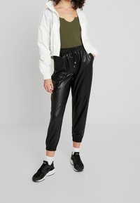 Weiße Zip-Jacke, olivfarbener Tanktop, schwarze Jogginghose aus Kunstleder mit elastischen Bündchen und schwarze Sneaker mit weißen Sohlen.
