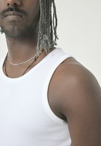White ribbed tank top with a round neckline, featuring a subtle embossed logo on the chest. Skin tone visible on the shoulder and neck.
