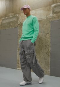 Sweatshirt verde menta, calças cargo cinzas com bolsos e ténis brancos. O modelo usa um boné rosa claro e está em um ambiente minimalista.