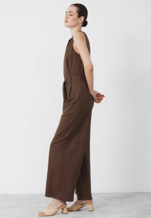 Kvinde står sideværts med lukkede øjne, iført en ærmeløs brun buksedragt med bælte og beige hælede sandaler mod en hvid baggrund.