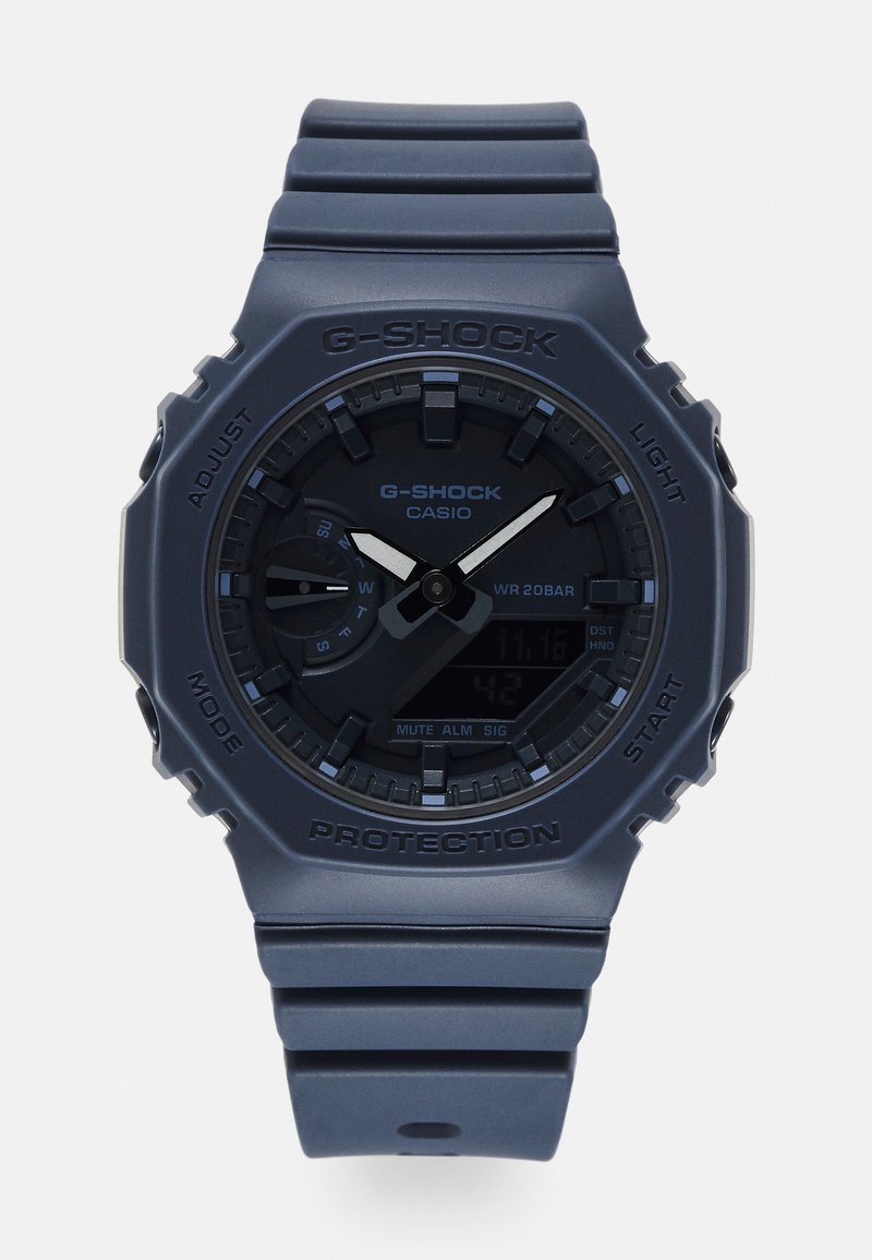 Analog-digital G-SHOCK klokke med et matt marineblått plasthus og rem. Har en svart skive med sølvdetaljer og flere funksjonsknapper.