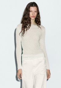 Jonge vrouw met lang golvend bruin haar, gekleed in een aansluitend wit textuur langsliepend bovenstuk en losse witte broek, staand voor een neutrale achtergrond.