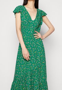 Grünes Blumenkleid mit V-Ausschnitt, schwingenden Ärmeln und schichtsaum. Der Stoff zeigt kleine rosa und gelbe Blumen.