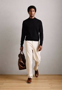 Un homme marchant à l'intérieur portant une chemise noire à manches longues, un pantalon crème, des chaussures marron et portant un sac en cuir marron.