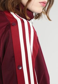 Maroon idrottsjacka med vita ränder, hög krage och tryckknappdetalj. Tillverkad av slät tyg med texturerad yta.