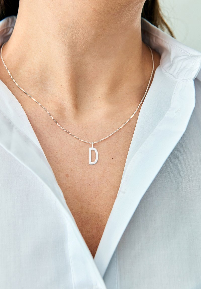 Silberne Kette mit einem Anhänger in Form eines großen "D", der an einer feinen Kette um einen nackten Hals hängt und vor einem hellblauen Hemd mit Kragen präsentiert wird.