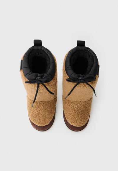 Bottes en polaire marron avec un revers noir rembourré, dotées d'un système de laçage et d'une semelle texturée, conçues pour le confort et la chaleur.