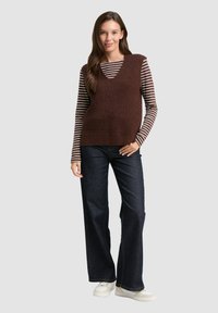 Eine Frau trägt eine braune gestrickte ärmellose Weste über einem braun-gestreiften Langarmshirt, dunklen weit geschnittenen Jeans und weißen Sneakers und steht.