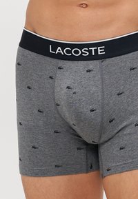 Boxer gris avec une ceinture élastique noire arborant "LACOSTE" en blanc. Décoré de petits logos crocodiles noirs.