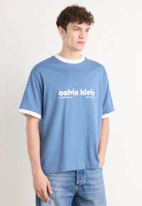 Blue cotton t-shirt with white ribbed collar and sleeve accents, featuring printed "calvin klein" logo and location text on the front.
