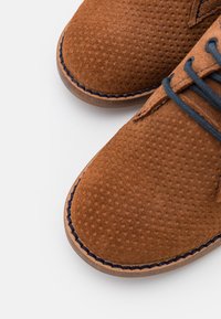 Hnědé semišové boty s texturovaným povrchem, kontrastním prošíváním a modrými tkaničkami. Kulatá špička a lehký design podešve.