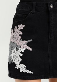Černá džínová sukně s květinovou výšivkou v růžové, šedé a bílé barvě. Obsahuje přední kapsy a zapínání na knoflík.
