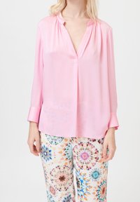 Femme portant un blouse rose loose à manches longues assortie à un pantalon blanc orné de motifs colorés en tie-dye circulaire.
