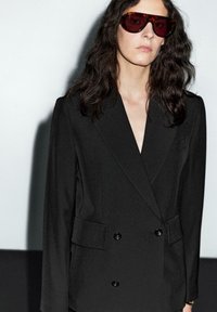 Černý dvouřadý blazer s klopovým límcem, se dvěma předními kapsami a černými knoflíky. Model nosí tmavé sluneční brýle s lesklým povrchem.