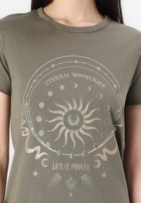 Oliivivihreä puuvillapaita, jossa on pyöreä graafinen kuva auringosta ja kuusta metallinhohtoisessa kullassa ja hopeassa, tekstillä "IKUISEN KUUNVALON" ja "KUUN VOIMA."
