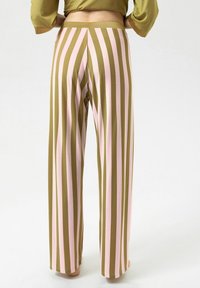 Persoon die een broek met hoge taille en wijde pijpen draagt, met verticale olijfgroene en lichtroze strepen, staand op blote voeten tegen een witte achtergrond.