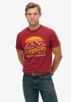 Red cotton t-shirt featuring graphic design with mountains and sun in yellow and orange. Paired with blue denim jeans and a brown belt.