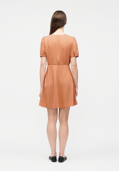 Γυναίκα που στέκεται γυρισμένη προς τα πίσω, φοράει ένα κοντομάνικο, μέχρι το γόνατο φόρεμα σε σκουριά απόχρωση και μαύρα παπούτσια χωρίς δέσιμο, μπροστά σε απλό λευκό φόντο.