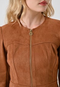 Vrouw in een bruine leren jas met een gouden rits en een decoratieve trekker, van schouders tot lippen gefotografeerd.
