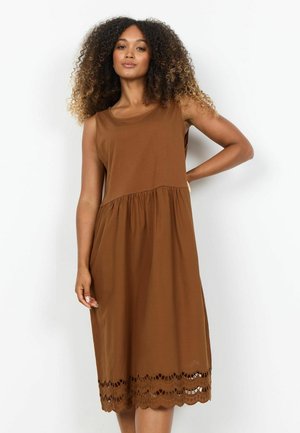 Kvinde med krøllet hår iført en ærmeløs brun midikjole med bølget blondekant, stående mod en hvid baggrund.
