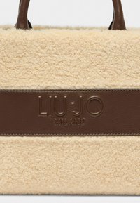 Sac fourre-tout en fausse fourrure crème avec une bandoulière en cuir marron et un accent logo. Texture douce, forme rectangulaire et détail de marque bien visible.