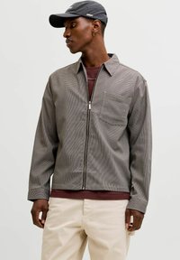 Graue kariert Reißverschlussjacke mit Kragen, einer Brusttasche und langen Ärmeln, getragen über einem dunkelroten Hemd und kombiniert mit beigen Hosen.