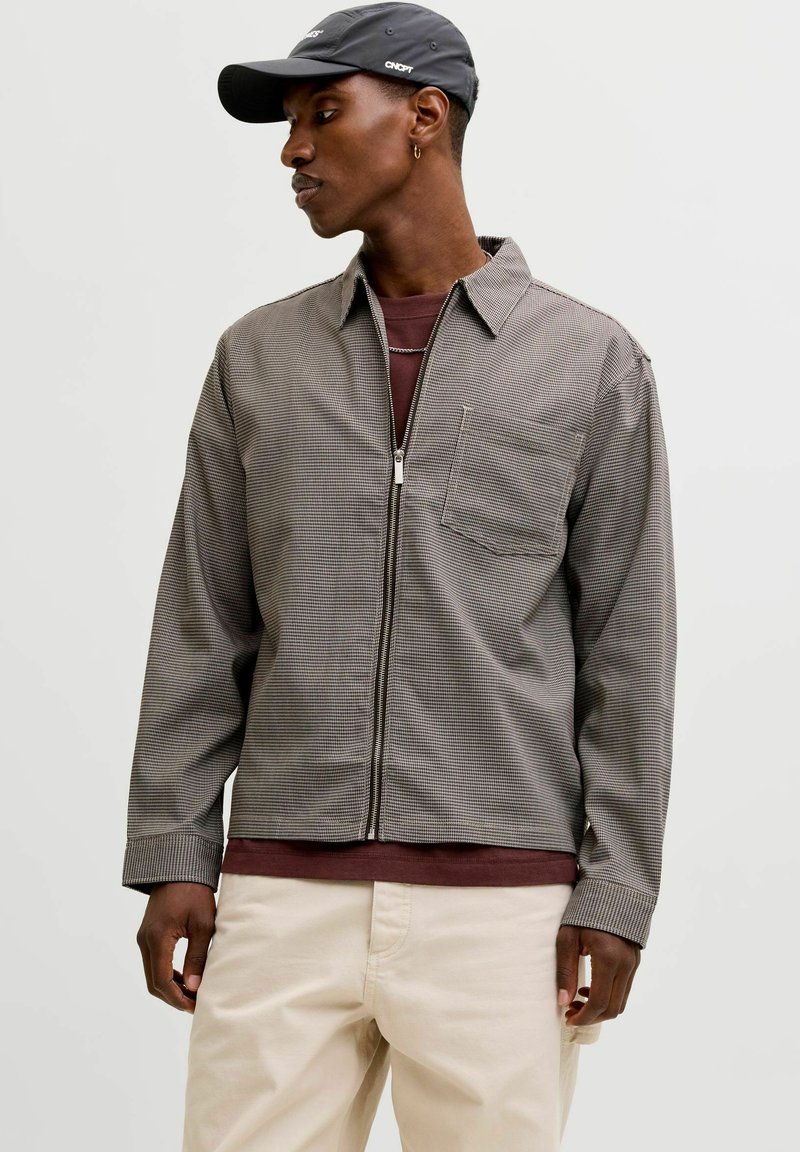 Graue kariert Reißverschlussjacke mit Kragen, einer Brusttasche und langen Ärmeln, getragen über einem dunkelroten Hemd und kombiniert mit beigen Hosen.