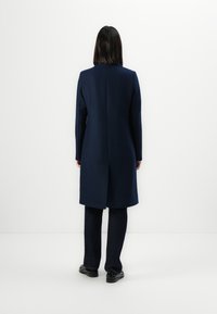 Marineblauer langer Mantel mit geradem Schnitt, langen Ärmeln und einer Mittelnaht hinten. Getragen über dunklen Jeans und schwarzen Schuhen.
