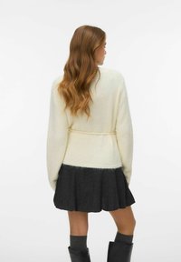 Roomkleurige gebreide trui met een tailleband, voorzien van een zachte textuur. Gecombineerd met een donkergrijze uitlopende rok, grijze kniehoge sokken en zwarte laarzen.