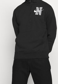 Black sweatshirt with a zippered collar, featuring a gray embroidered "N" logo and Nike swoosh. Soft fabric with a front kangaroo pocket.