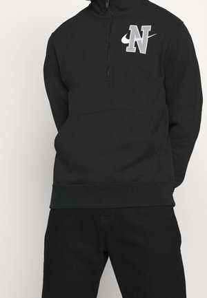 Sudadera negra con cuello con cremallera, que presenta un logo "N" bordado en gris y el swoosh de Nike. Tejido suave con un bolsillo tipo canguro en la parte delantera.