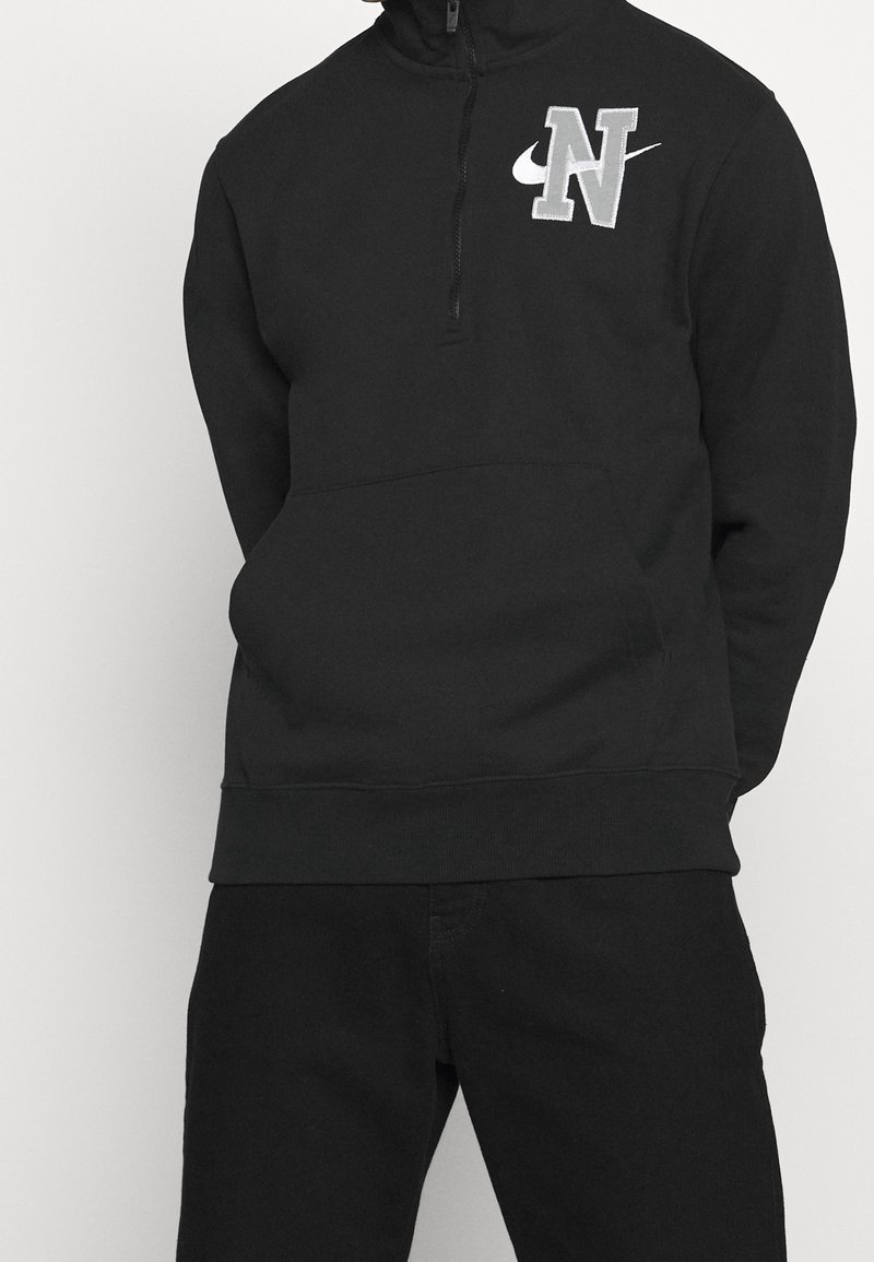 Black sweatshirt with a zippered collar, featuring a gray embroidered "N" logo and Nike swoosh. Soft fabric with a front kangaroo pocket.