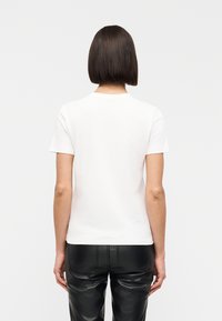 Γυναίκα με ίσια, μαύρα μαλλιά μέχρι το πιγούνι, που φοράει απλό λευκό κοντομάνικο πουκάμισο και μαύρο δερμάτινο παντελόνι, που φαίνεται από πίσω.