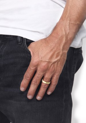 Gold-colored metal ring worn on a hand with a textured, creased skin tone, resting on dark denim jeans. Simple, smooth design.