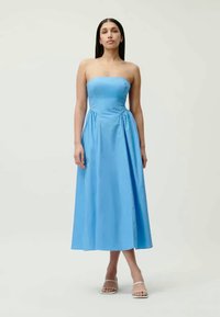 Ένα μεσαίου μήκους, strapless φόρεμα από ανοιχτό μπλε ύφασμα, με εφαρμοστό μπούστο και μαζεμένα λεπτομέρειες στη μέση, συνδυασμένο με λεπτές παντόφλες.