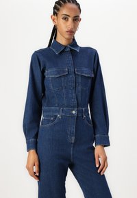 Denim jumpsuit met lange mouwen, kraag, twee borstzakken en een getailleerde taille. Donkerblauwe kleur met zichtbare stikseldetails.