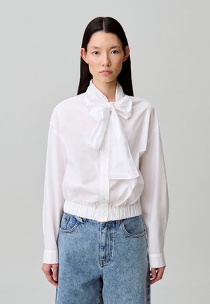 Chemise blanche boutonnée avec col à cravate, taille élastiquée et manches longues. Portée avec un short en denim taille haute. Texture lisse.