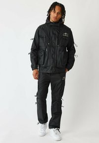 Zwarte waterafstotende jas met meerdere zakken en aansnoerdetails, gecombineerd met bijpassende zwarte broek en witte sneakers.
