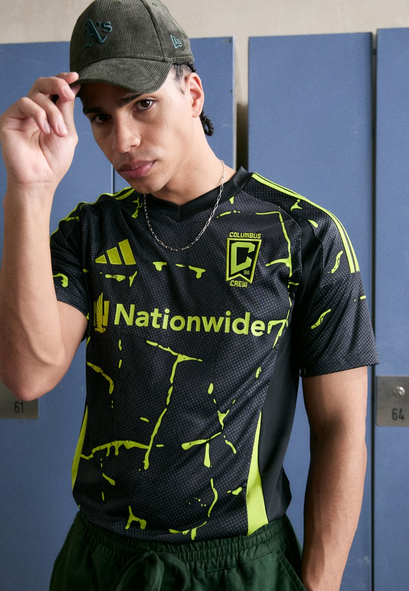 Jersey negra con diseño de salpicaduras en verde neón y detalles. Presenta un escote en forma de V, logo de Adidas y logo de "Nationwide". Gorra gris completa el look.