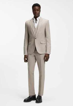 Homme portant un costume beige deux pièces avec une chemise blanche et des chaussures habillées noires, debout devant un fond blanc uni.