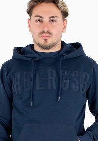 Felpa con cappuccio di colore blu navy realizzata in tessuto morbido, dotata di un grande logo impresso sul davanti e di una tasca a canguro. Inclusa anche la coulisse per il cappuccio.