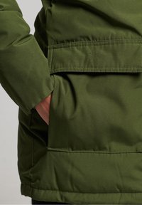 Hand in de zak van een groene gewatteerde jas met een grote klepzak aan de voorkant en zichtbare stiksel details.