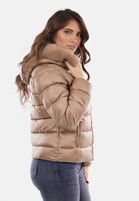 Giacca a vento marrone chiaro con cappuccio foderato di pelliccia, caratterizzata da sezioni trapuntate orizzontalmente e chiusura con zip. Tessuto liscio e lucido.