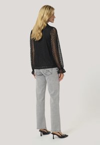 Svart spetsblus med geometriska mönster, långa genomskinliga ärmar och volangkant; i kombination med ljusgrå högmidjade jeans och svarta klackmules.