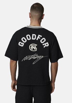 Homme aux cheveux courts portant une chemise noire à manches courtes texturée avec l'inscription "GOODFOR nothing" et un logo circulaire dans le dos, debout devant un fond blanc.